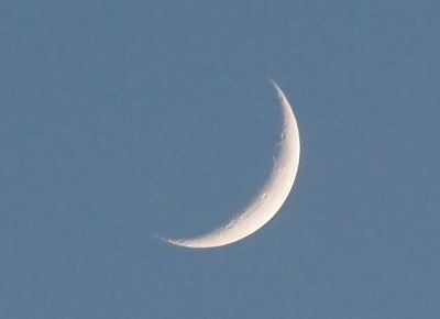 [The early night sky is light blue. A lower right sliver of moon is visible with a sharp outer edge. The inner edge is a more subtle color change and there are several darker round sections in the white which are moon craters.]